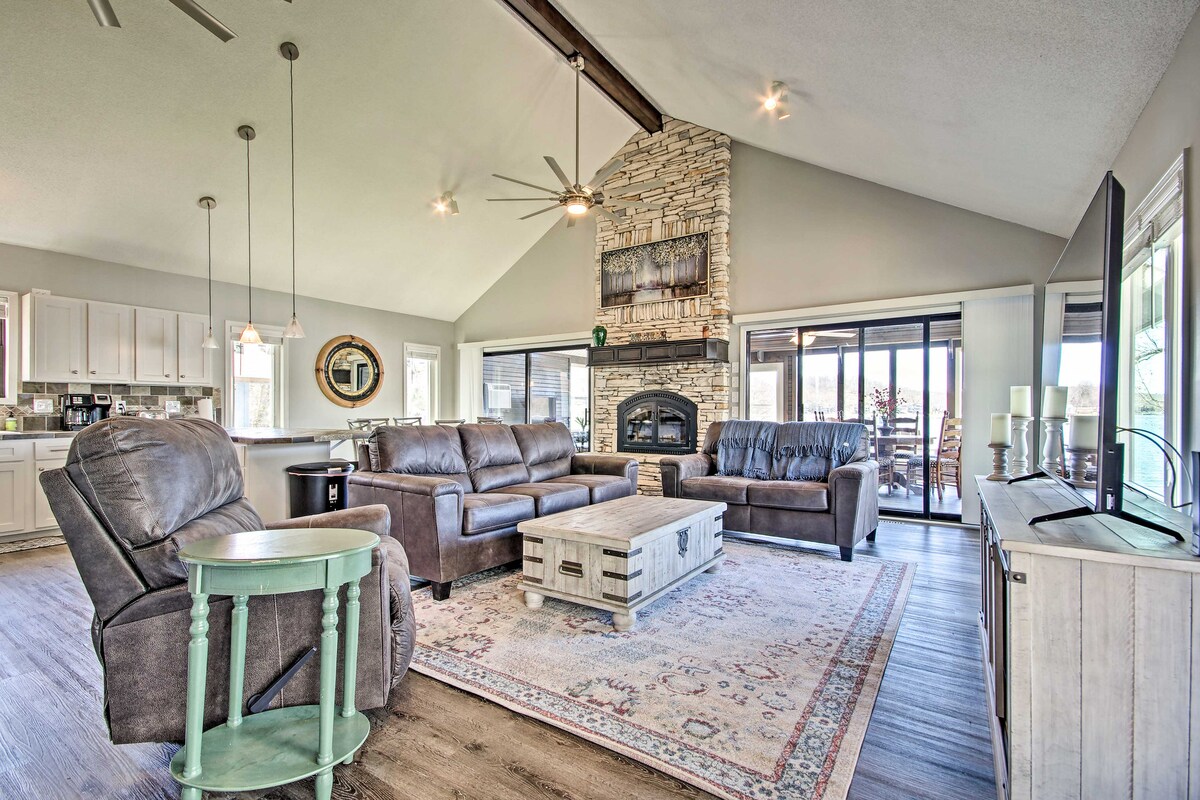 The living area features high ceilings with ceiling fans and natural light from large windows. A stone fireplace serves as a focal point, complemented by comfortable leather seating. A rustic coffee table is placed on a patterned area rug, with a view of the outdoor deck visible through sliding doors.