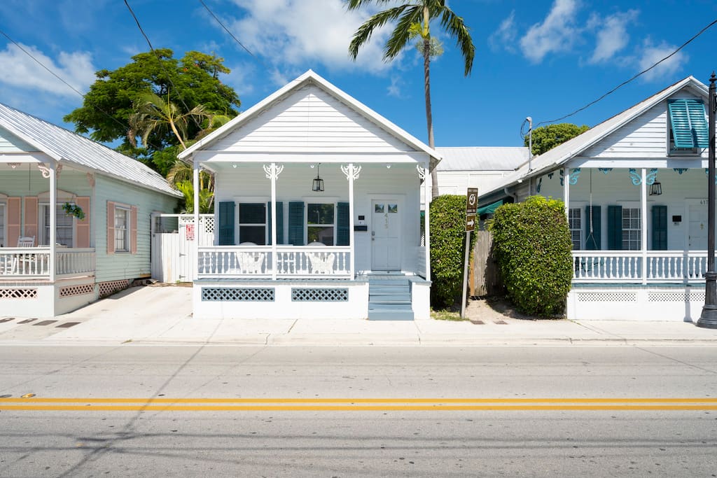 The exterior of a charming white cottage is visible, complete with a welcoming porch and blue accents. Lush greenery surrounds the property, and palm trees rise toward a bright blue sky. The neighboring cottages exhibit a similar architectural style, enhancing the inviting atmosphere of the street.