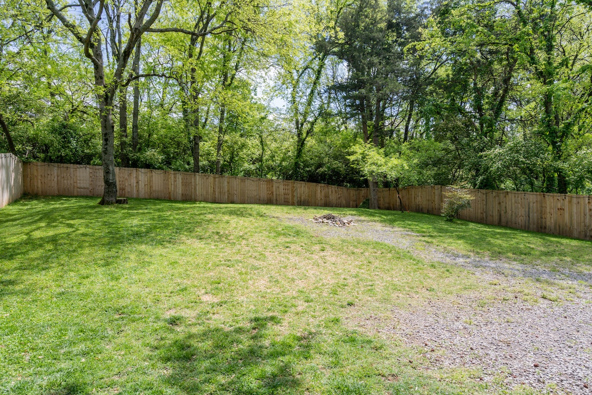 A spacious fully fenced-in backyard is presented, featuring a grassy area surrounded by trees. A gravel path leads to a small fire pit, providing a secure space for outdoor activities. The wooden fence offers privacy while maintaining a natural setting.