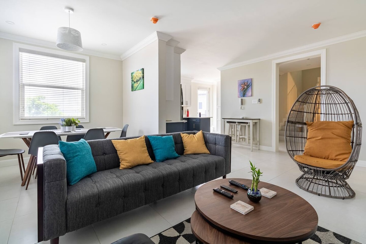 An open living area is presented with a dark gray sofa adorned with blue and yellow cushions. A round wooden coffee table sits in front, surrounded by a patterned rug. An elegant dining area features a light-colored table and chairs, while a wicker chair adds a touch of comfort.