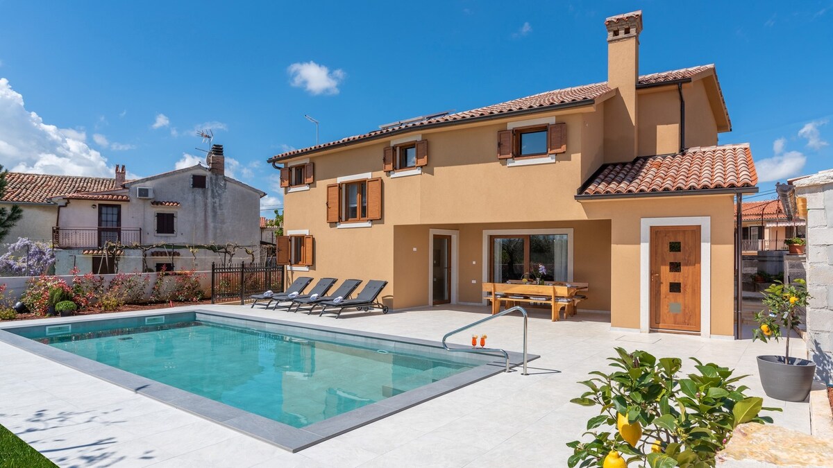 The exterior of a modern villa showcases a spacious pool surrounded by sun loungers. The building features large windows and an inviting outdoor dining area. A well-maintained garden with greenery enhances the serene atmosphere of the property.