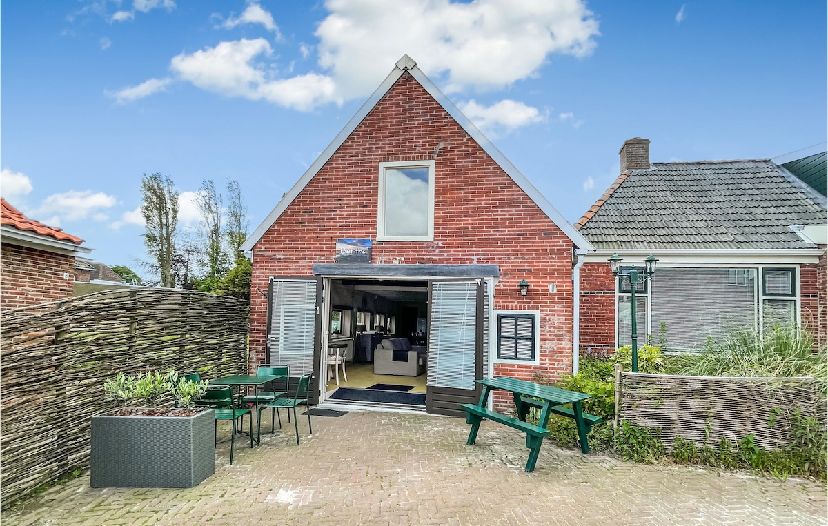 The exterior of the holiday home is shown, featuring a red-brick façade and large glass doors that open to the living space. Outdoor seating is visible in the foreground with green chairs and a planter. Lush greenery surrounds the area, contributing to a welcoming atmosphere.