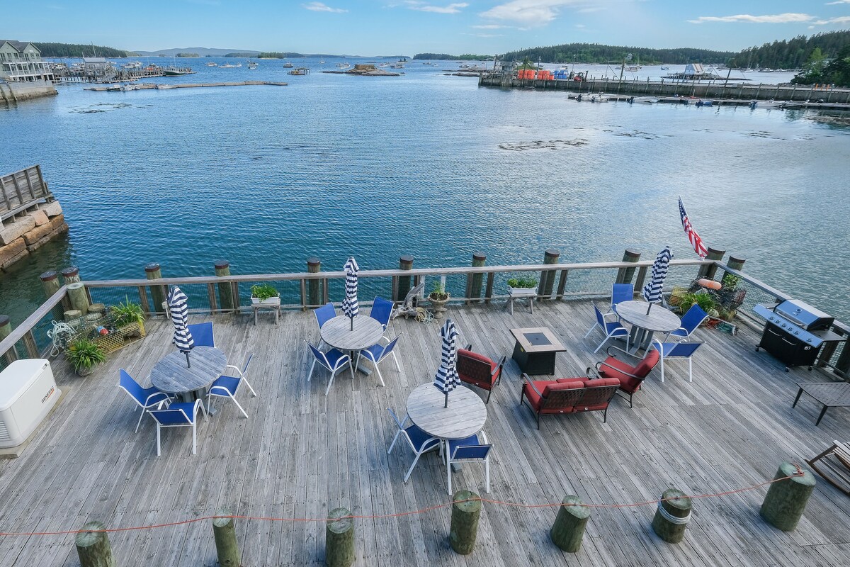 An expansive deck overlooks the harbor, featuring several round tables and blue chairs, some with striped umbrellas. A fire pit is centered among the seating arrangements, and a barbecue grill is visible on one side of the deck, creating a welcoming outdoor space.