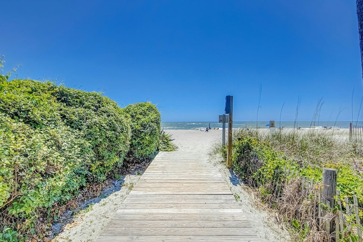 A wooden boardwalk leads through lush greenery to the sandy beach, which gently meets the ocean. The bright blue sky adds to the vibrant atmosphere, creating an inviting access point to the shore.