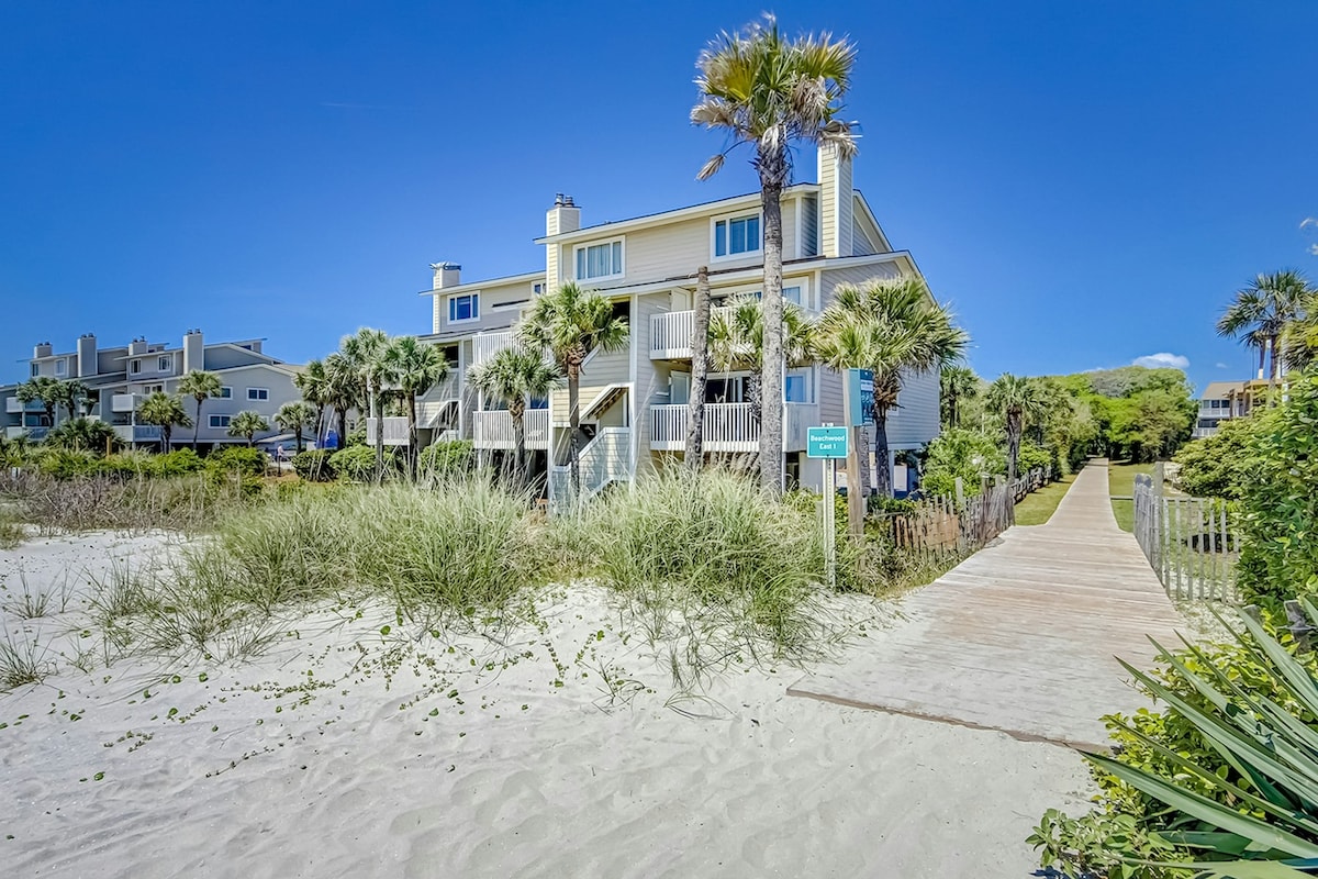 The exterior of a beachside villa is visible, situated among palm trees and beach grass. A sandy path leads to the entrance of the property, with wooden walkways providing access to the sandy shore. Clear blue skies provide a sunny backdrop.