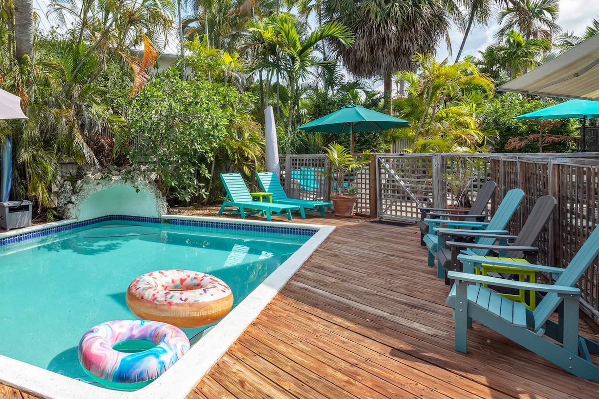 A tropical backyard features a shared pool surrounded by a wooden deck. Colorful lounge chairs are arranged for relaxation, while umbrellas provide shaded seating areas. Two inflatable pool floats rest in the water, adding a playful touch to the serene, lush landscape.