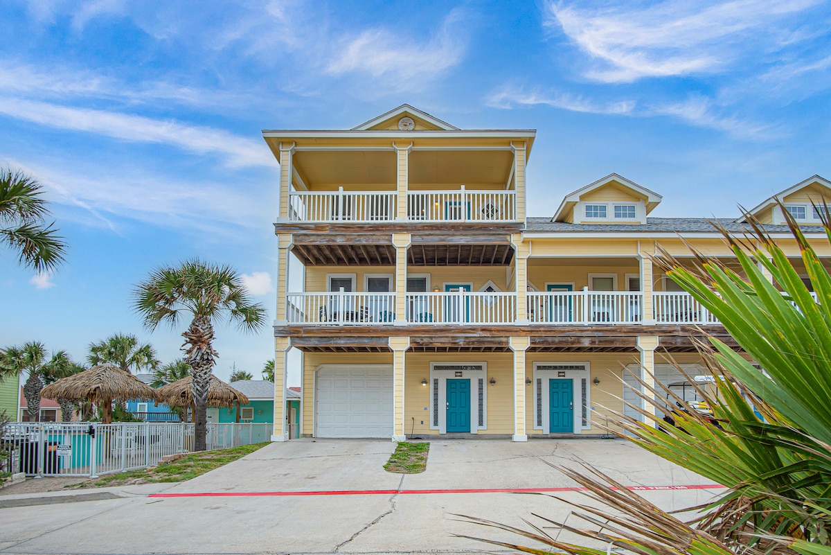 The two-story townhome features a welcoming yellow exterior with white railings. A spacious garage is visible on the ground level, flanked by palm trees. Two large balconies are situated on the second floor, providing ample outdoor space, with a palapa-style seating area in the background.