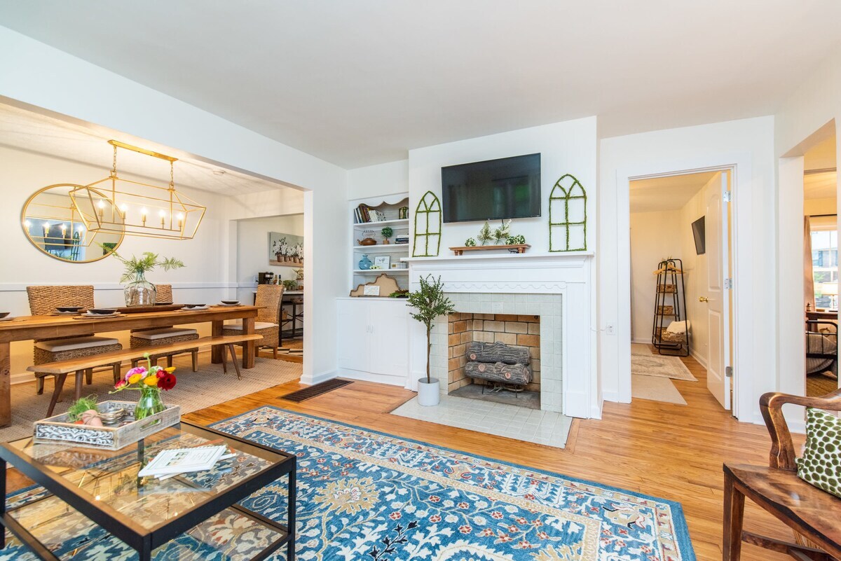 A bright living area features a white brick fireplace with a decorative mantel. A large television is mounted above the fireplace. A coffee table with floral arrangements is centered on a patterned area rug, while a dining area with a long table and woven chairs is visible in the background.