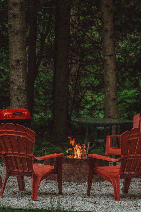 A fire pit surrounded by four red Muskoka chairs is highlighted in a secluded outdoor setting. A BBQ grill is positioned nearby, with lush greenery in the background, creating a space ideal for relaxation and gatherings.