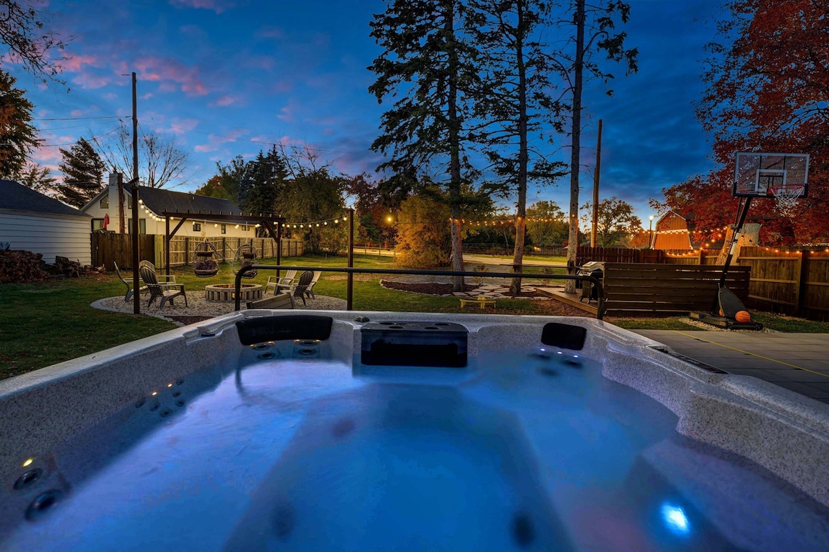 A spacious hot tub is illuminated in soft blue light, surrounded by a well-maintained backyard. The area features a basketball hoop, a cozy seating arrangement, and string lights in the trees, creating a serene setting at dusk.