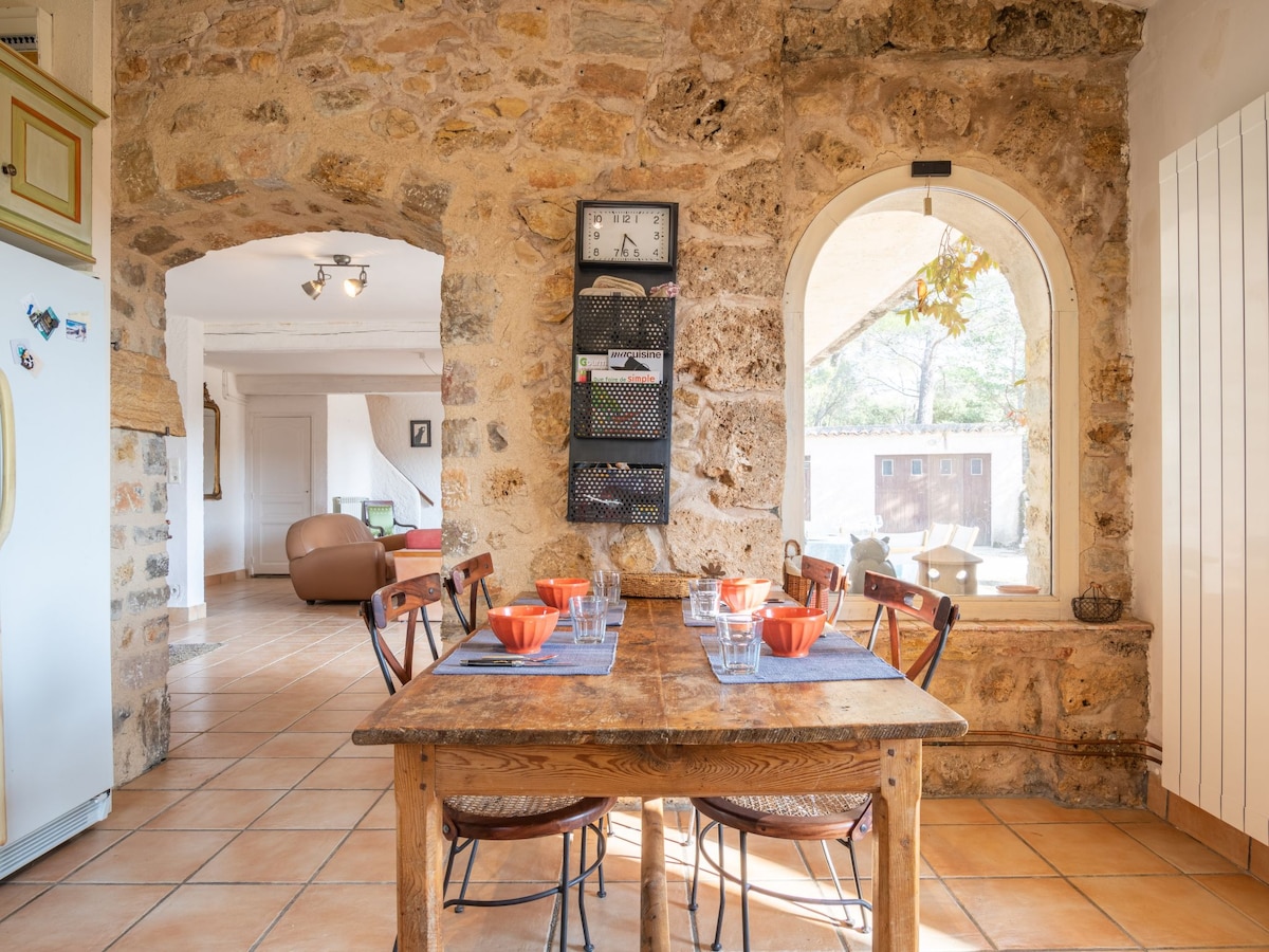 A rustic dining area features a wooden table set for six, with colorful bowls arranged beside glasses. The stone walls and arched window provide a warm, natural backdrop, while soft light enters from outside, enhancing the space's inviting feel.