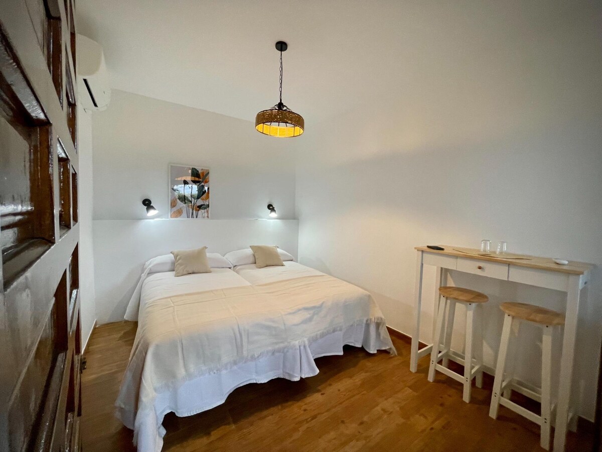 A bright sleeping area features two spacious beds covered in white linens. The room is complemented by contemporary bedside lamps and a small bar table with two stools. Light wood flooring adds warmth, while a decorative piece hangs on the wall.