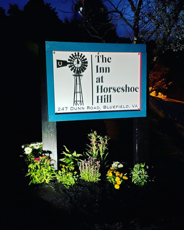 A welcoming sign for The Inn at Horseshoe Hill is displayed prominently at night, illuminated by soft lighting. The sign features a windmill design and includes the address, with a surrounding garden of blooming flowers and shrubs adding a touch of natural beauty.