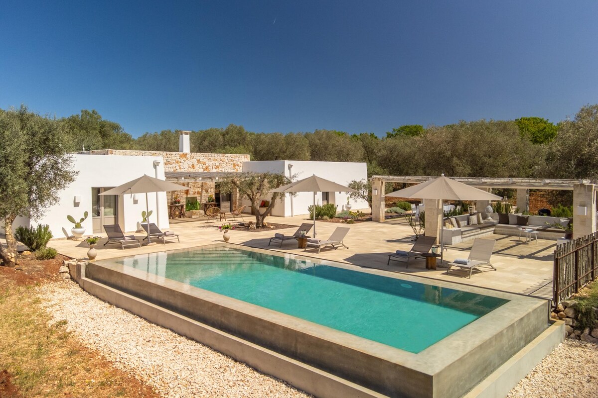 An inviting outdoor space features an infinity pool surrounded by sun loungers and umbrellas. Olive trees provide shade, while a spacious patio includes an outdoor dining area and seating arrangements. The residence is characterized by a modern aesthetic with light-colored walls.