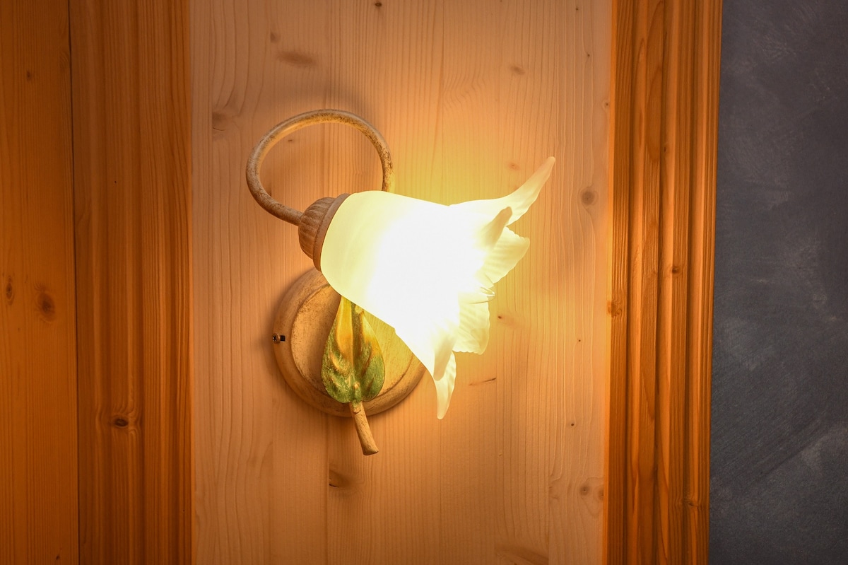 A wall-mounted light fixture designed in the shape of a flower, featuring a soft, glowing bulb surrounded by a delicate glass petal. The fixture is positioned against warm wooden paneling, enhancing the natural ambiance of the space.