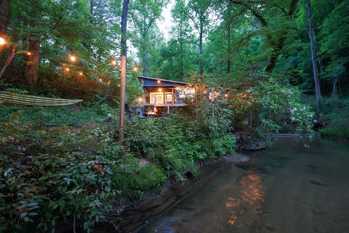 The cabin is nestled amidst lush greenery, illuminated by outdoor string lights. A creek runs beside the property, which features a small deck area overlooking the water. Surrounding trees add to the tranquil, woodland atmosphere.
