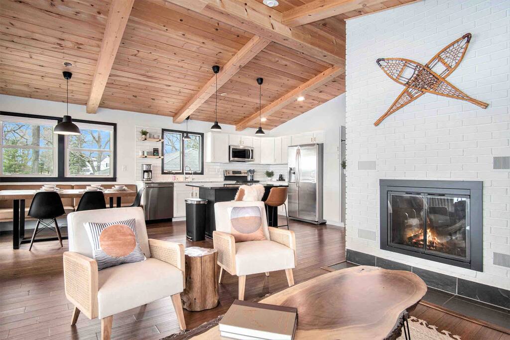 A bright and open living area features a vaulted wooden ceiling and large windows providing natural light. A modern kitchen is visible in the background, alongside a dining table. Two comfortable chairs are positioned near a gas fireplace, creating a relaxing space.