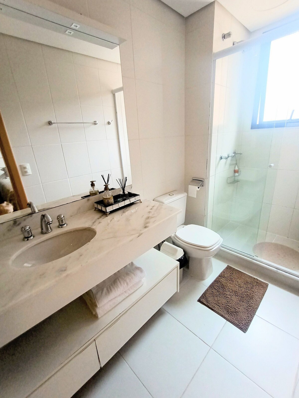 A modern bathroom features a marble countertop with a sink and a decorative arrangement of toiletries. A glass shower enclosure is visible, alongside a toilet and woven bath mat. Natural light enters through a window above the shower, enhancing the space's clean aesthetic.