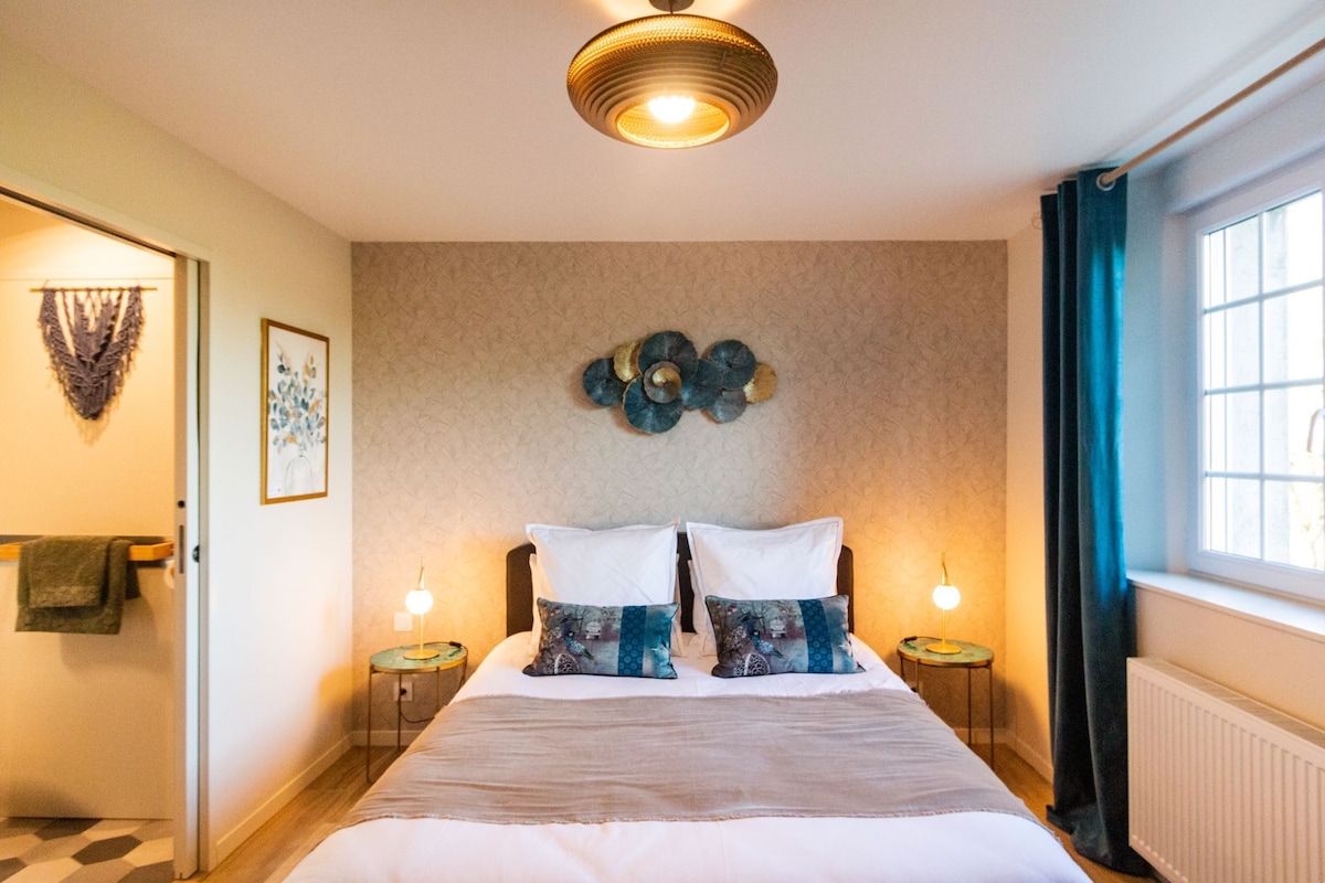 A spacious bedroom is presented with a large bed adorned with a soft blanket and decorative pillows. Elegant bedside lamps are positioned on either side. Natural light enters through a window dressed with sheer curtains, complemented by a tasteful wall decoration featuring shades of blue and gold.