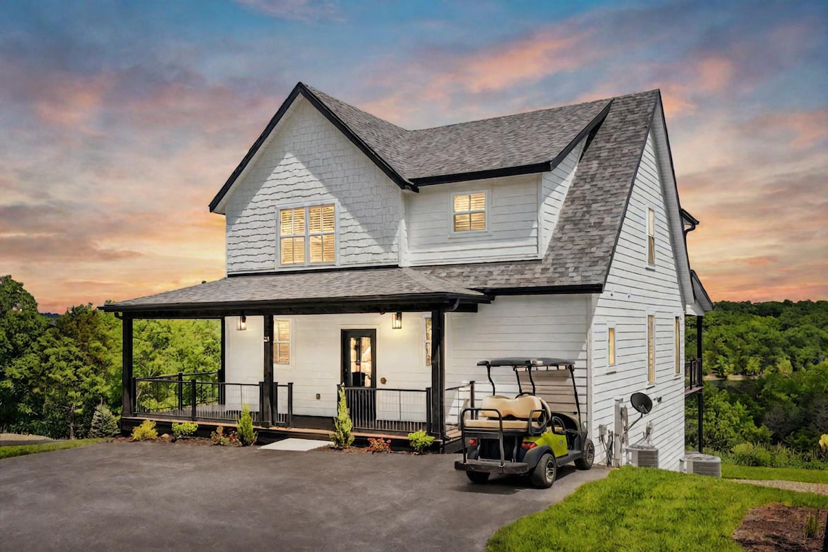 A modern farmhouse exterior features a two-story structure with a welcoming porch and large windows. The driveway is lined with neatly arranged landscaping, and a golf cart is parked nearby, showcasing the property’s recreational amenities. The sky transitions with soft hues as the sun sets.