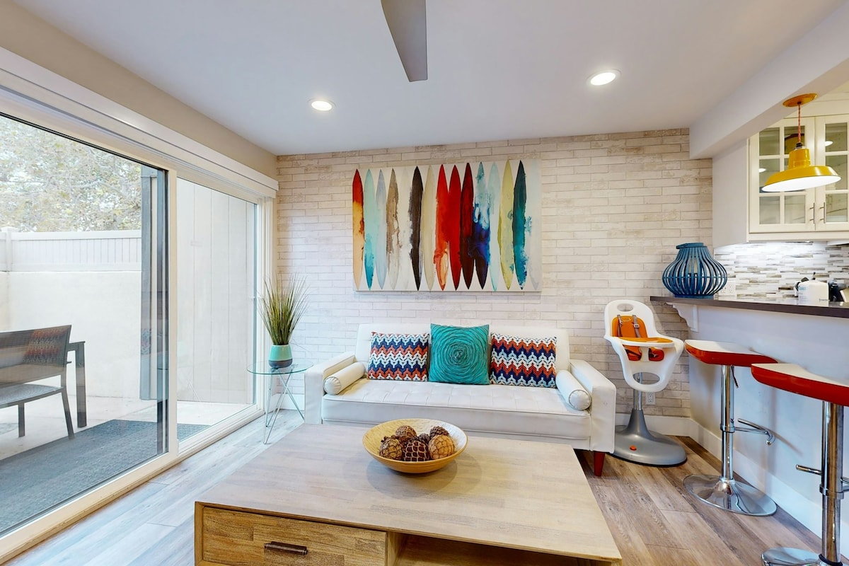 A bright living area features a white couch adorned with colorful pillows, complemented by a wooden coffee table displaying decorative bowls. A large artwork featuring surfboards adds color to the light brick wall. Sliding glass doors lead to an outdoor space, enhancing natural light.