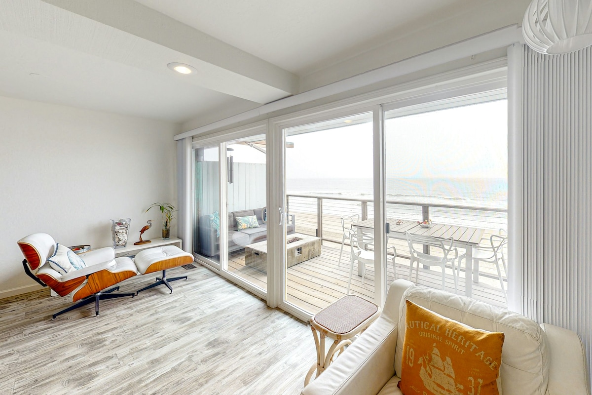 An open living area features large sliding glass doors that provide natural light and stunning ocean views. Comfortable seating options are available, including a modern lounge chair and a cozy sofa. A dining space is positioned outside on the deck, inviting a seamless indoor-outdoor experience.