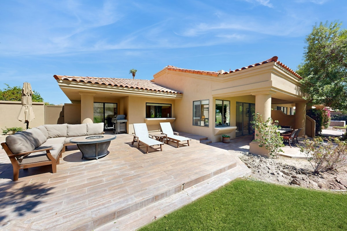 A spacious outdoor area features a comfortable sofa surrounding a gas firepit. Two sun loungers are positioned nearby, and a gas grill is visible to the side. Lush landscaping contributes to a serene environment under a clear blue sky.