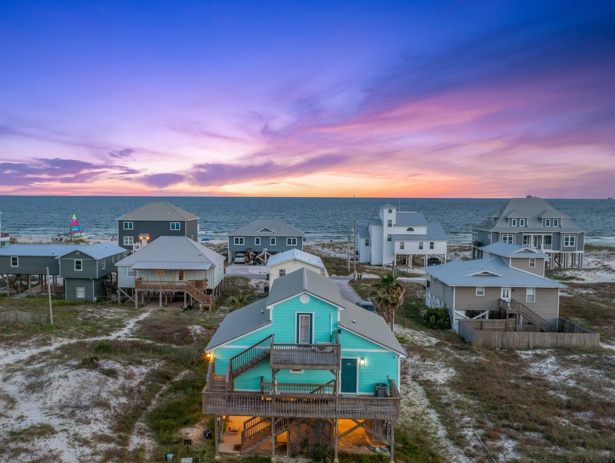 A view of several beach homes is highlighted under a vibrant sunset sky, showcasing rich hues of pink and purple. The sandy landscape reflects the coastal environment, while the homes feature varying architectural styles and colors, creating a charming coastal community.
