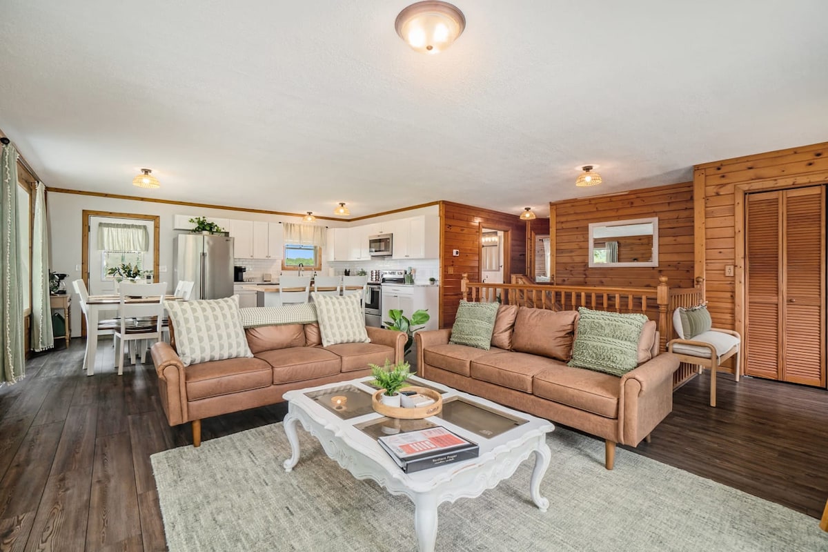An open-concept living area showcases a cozy seating arrangement with neutral-toned sofas and a layered coffee table. Natural light fills the space, highlighting the wooden accents and modern kitchen visible in the background. A dining area with a table for six is situated towards the rear.