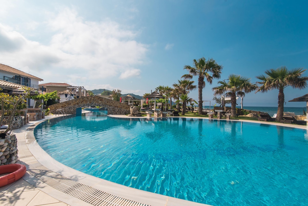 A large outdoor pool is framed by palm trees and lounge areas, capturing a serene atmosphere. The water reflects the clear blue sky, while stone pathways provide access to nearby seating and shaded structures. The ocean can be glimpsed in the background.