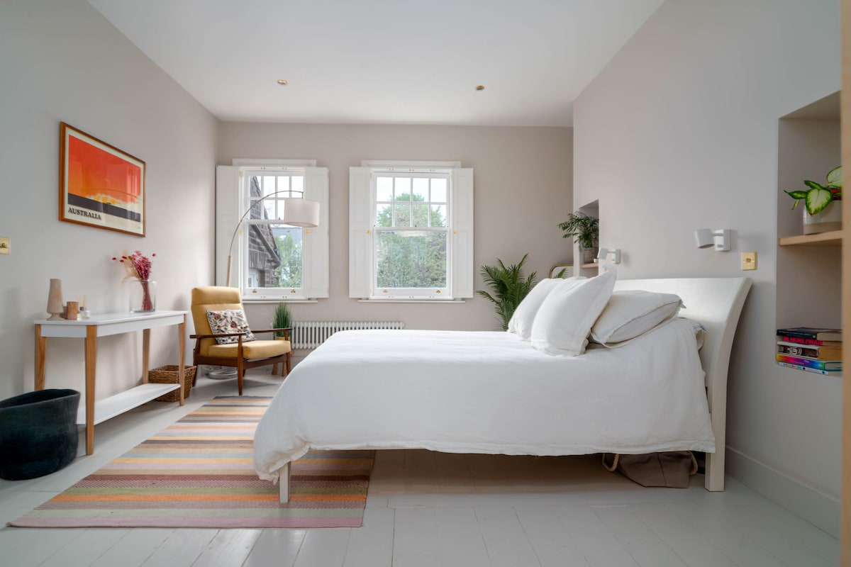A light and spacious bedroom features a large bed with soft white bedding. Two windows allow natural light to fill the room, illuminating the neutral-colored walls. A small desk and chair are positioned nearby, alongside a plant and colorful area rug on the floor.