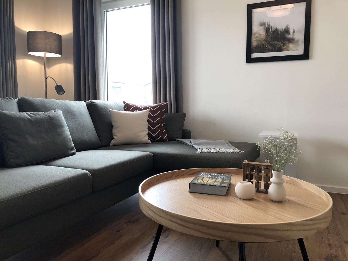 A modern living area features a plush corner sofa adorned with decorative cushions. A wooden coffee table, centered in the room, displays a book and small decor items. Natural light enters through a window, framed by flowing curtains, enhancing the cozy atmosphere.