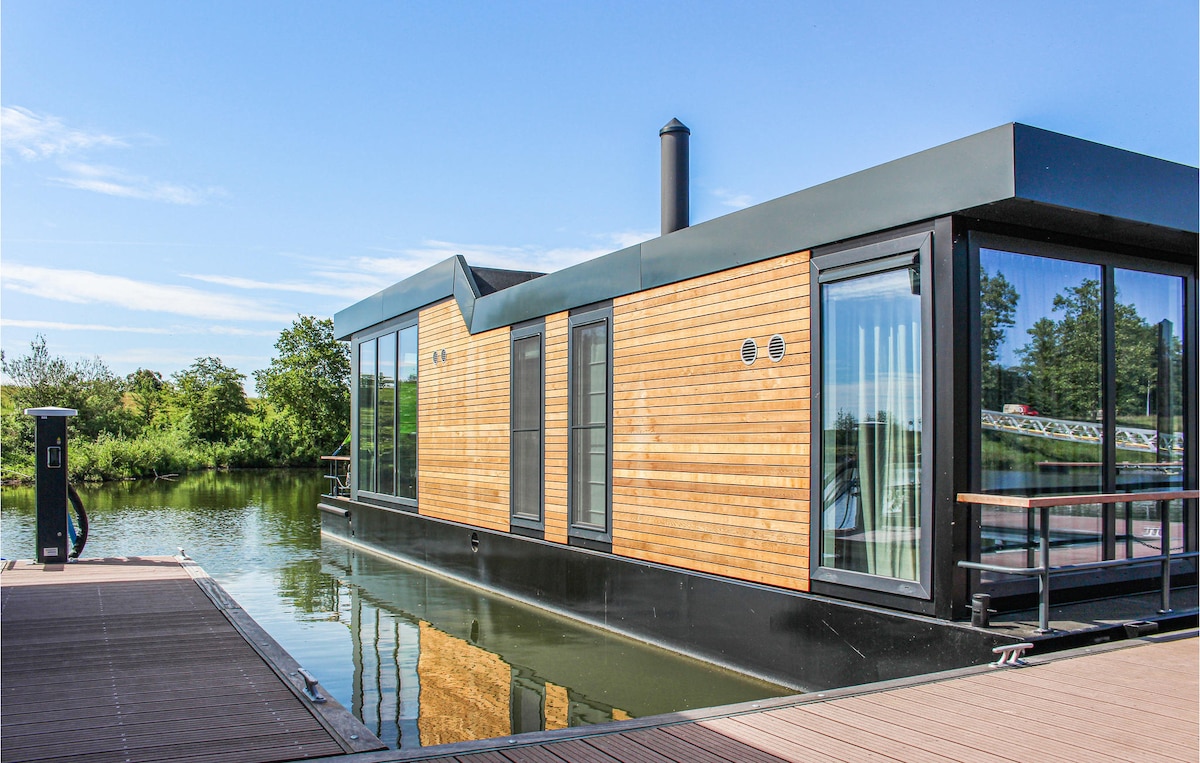 A modern houseboat is positioned on calm waters, featuring a timber exterior and large glass panels. A terrace extends outward, providing direct access to the water. Surrounding greenery enhances the tranquil setting, and a clear blue sky is visible above.