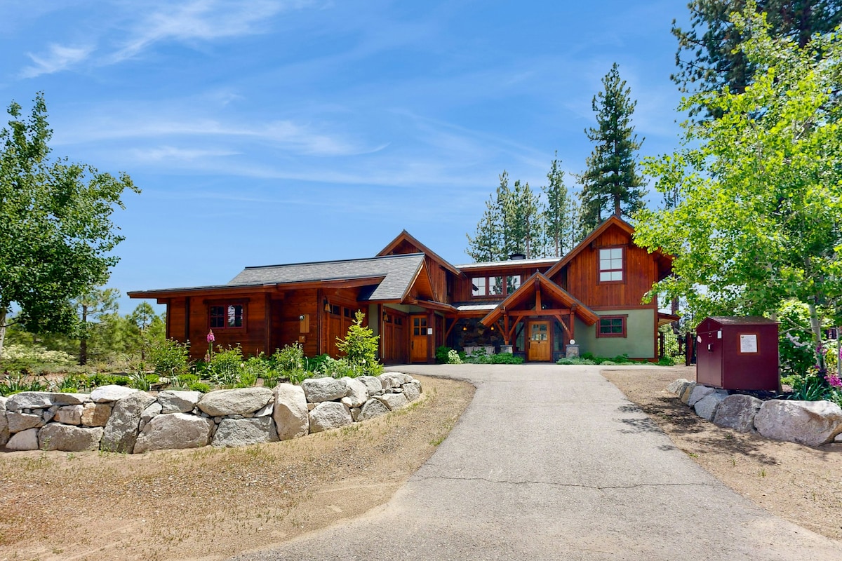 A well-crafted, multi-wing home is surrounded by lush greenery and tall trees. A paved driveway leads up to the welcoming entrance, framed by neatly maintained landscaping and rock features.