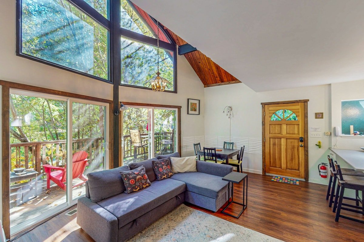 The living area features vaulted ceilings and large windows allowing natural light to fill the space. A comfortable sofa and armchair are positioned around a side table, with a dining area for four visible in the background. An inviting entrance door adds warmth to the setting.