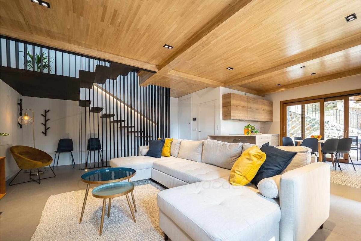 A bright living area is highlighted by a spacious sectional sofa adorned with yellow pillows. A round glass coffee table sits on a soft area rug. Natural light filters through large glass doors, revealing an inviting outdoor space. A modern staircase adds to the contemporary feel.