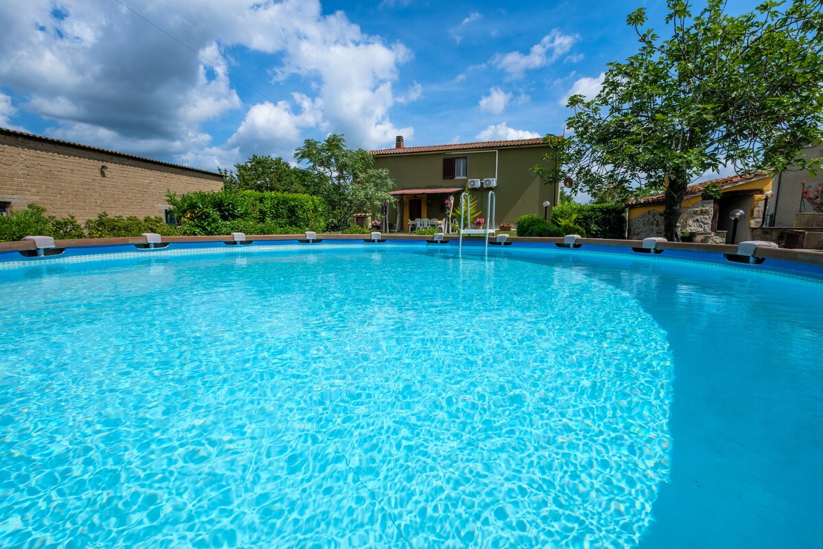 The above-ground swimming pool, with a 5 m diameter, features clear blue water surrounded by a paved area with lounging chairs. Green foliage and a two-story holiday home can be seen in the background, under a bright sky dotted with clouds.