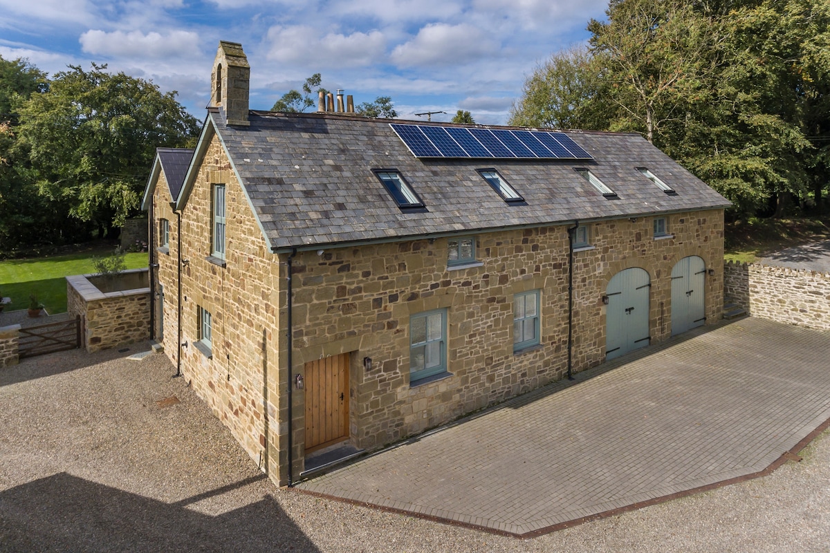The exterior of Glaneirw Coach House showcases a beautifully restored stone façade with a slate roof and solar panels. The building features large, light green doors and a wooden entrance, surrounded by a gravel driveway. Lush greenery is visible in the background.