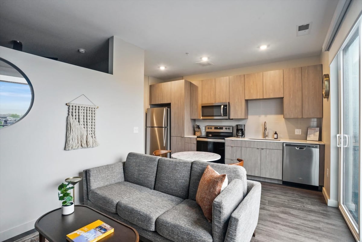 An open-concept living area features a modern grey sofa with an accent pillow, a circular dining table, and sleek kitchen cabinetry. Stainless steel appliances are visible in the kitchen, while natural light flows in from a large sliding glass door, enhancing the inviting atmosphere.