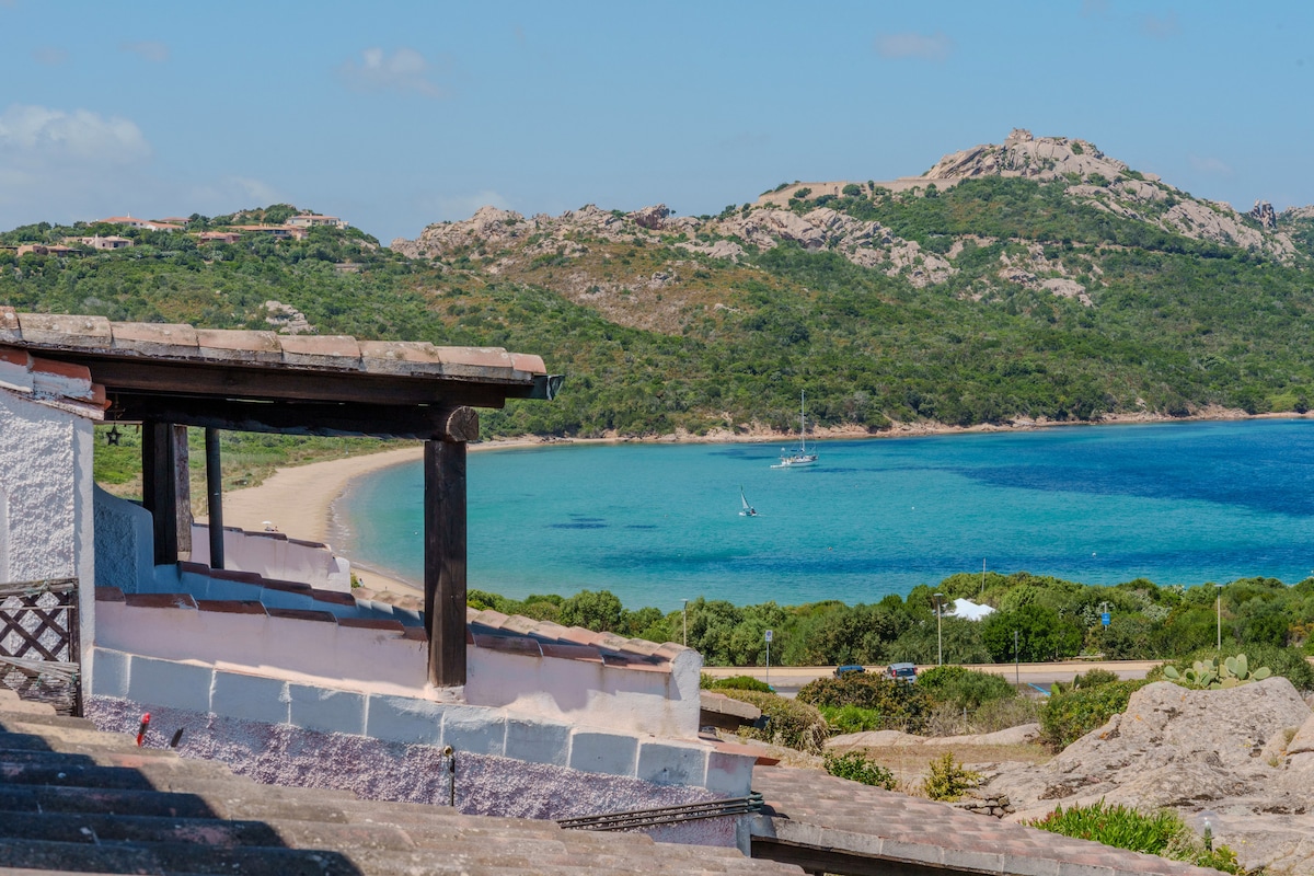 A panoramic view captures the serene coastline, featuring a sandy beach stretching along calm turquoise waters. Verdant hills rise in the background, and a sailboat can be seen gently bobbing on the water. The image highlights the natural beauty surrounding the residence.