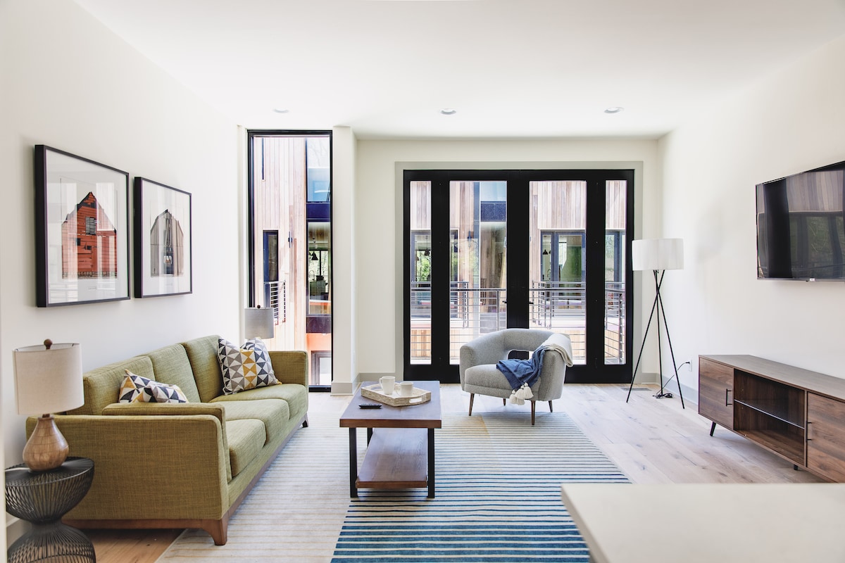 A bright living area features large glass doors opening to a patio, allowing natural light to fill the space. A green couch, a lounge chair, and an accent table create a comfortable seating arrangement. Framed artwork adorns the walls, and a TV is mounted on the opposite side.