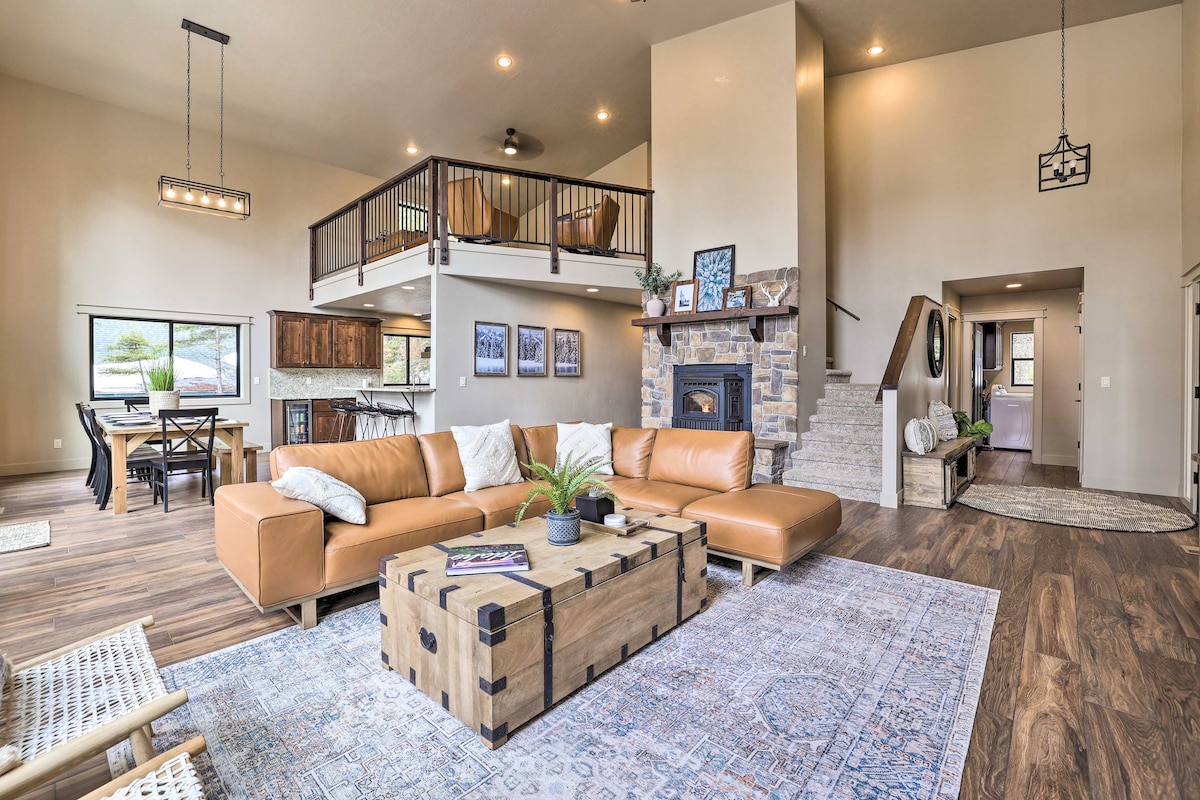 The spacious living area features a modern leather sectional sofa arranged around a wooden coffee table. A cozy wood pellet fireplace is set into a stone wall, and large windows allow natural light to fill the room. A dining area and staircase to a loft can be seen in the background.