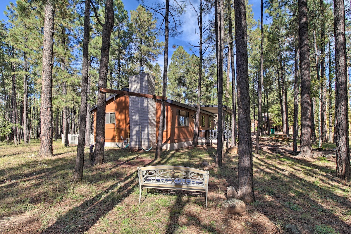 A serene cabin surrounded by tall pine trees, offering a peaceful setting. The structure features a stone chimney and a covered porch, with a bench positioned in the foreground, inviting guests to enjoy the natural surroundings.