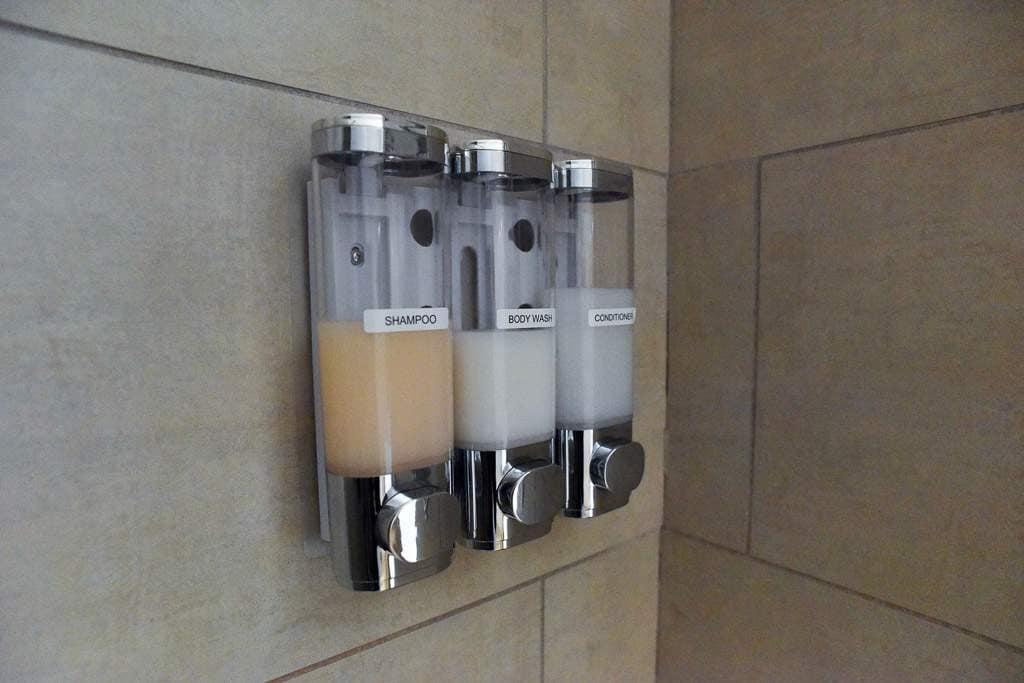 Three dispensers are mounted on the shower wall, containing shampoo, body wash, and conditioner. Each dispenser is labeled and has a clear design, showcasing the contents. The surrounding tiles feature a neutral color palette, enhancing the modern bathroom aesthetic.