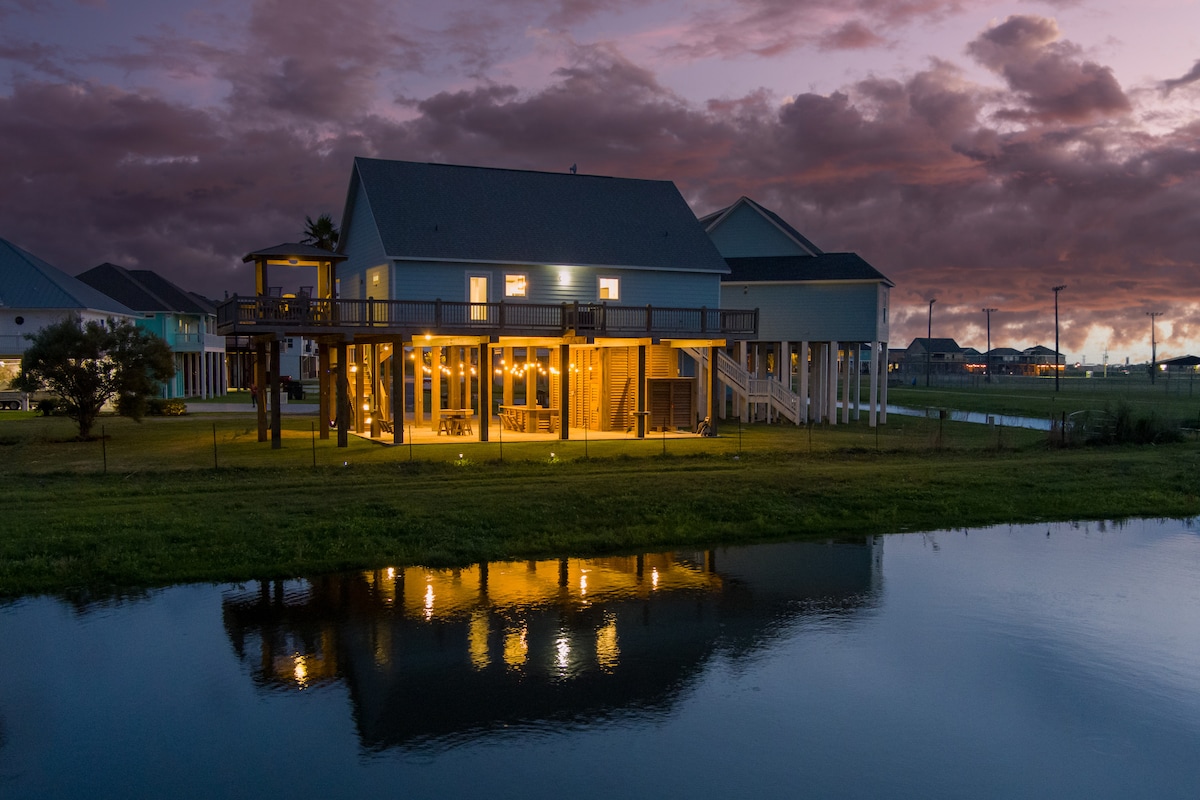 A spacious home features large windows showcasing illuminated interiors against a backdrop of a dramatic sunset. The structure is positioned near a serene water body, with reflections of the home visible on the calm surface. The surrounding landscape includes well-maintained grass and distant properties.