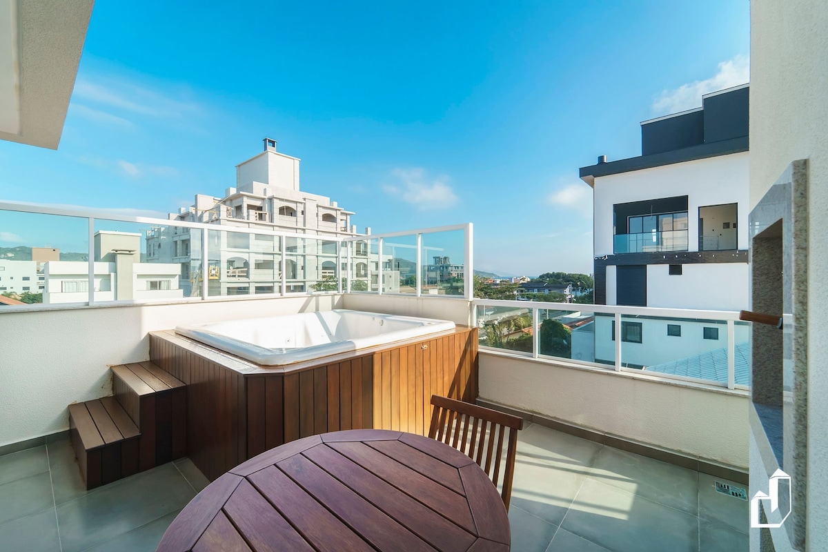 An outdoor terrace features a spacious wooden jacuzzi accompanied by a round table and a wooden chair. The area is surrounded by glass railings, offering clear views of the surrounding buildings and sky.