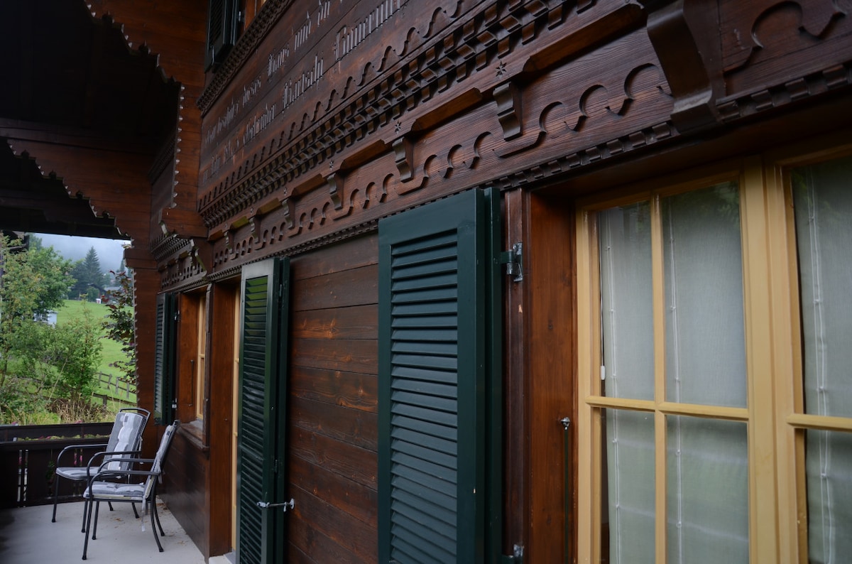 The wooden exterior of Chalet Finkenwiese features intricate carvings and shutters in shades of green. Two chairs are positioned on the porch, offering a space to relax and enjoy the surrounding landscape. The windows display soft yellow frames, enhancing the rustic charm.