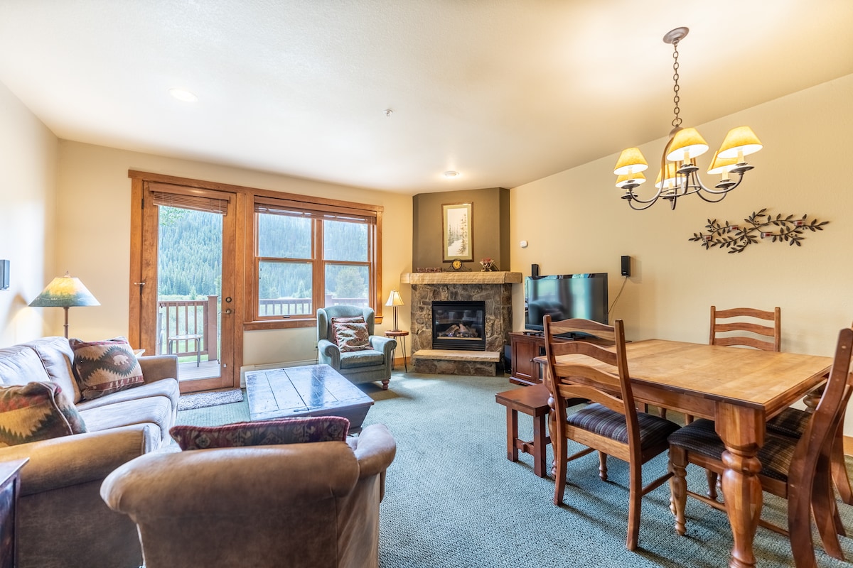The living area features a cozy seating arrangement with a sofa, armchair, and a coffee table. A gas fireplace is positioned next to the flat-screen TV. Dining is provided for six at a wooden table, with natural light streaming through double doors leading to the outdoors.