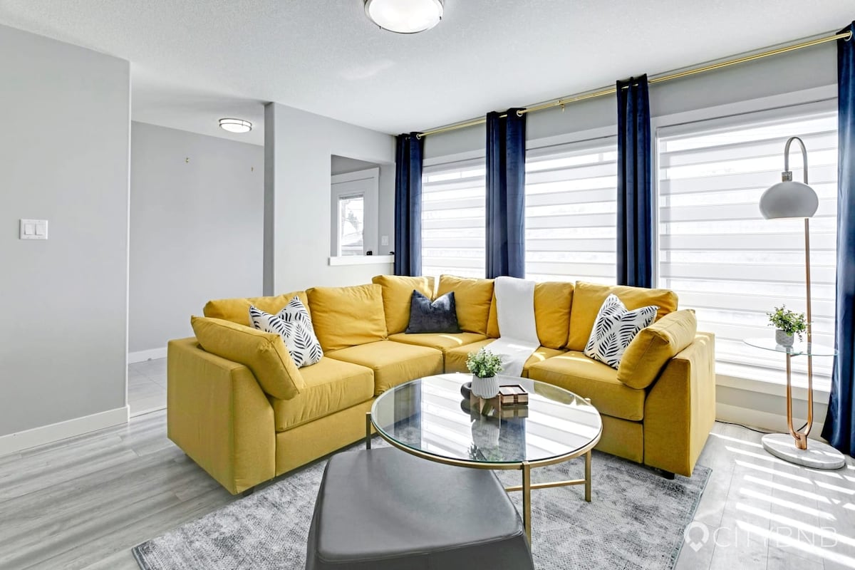 A spacious living area features a modern yellow sectional sofa with decorative cushions. A glass coffee table sits centrally, complemented by a small potted plant. Large windows adorned with sheer curtains allow natural light to fill the room, enhancing the inviting space.