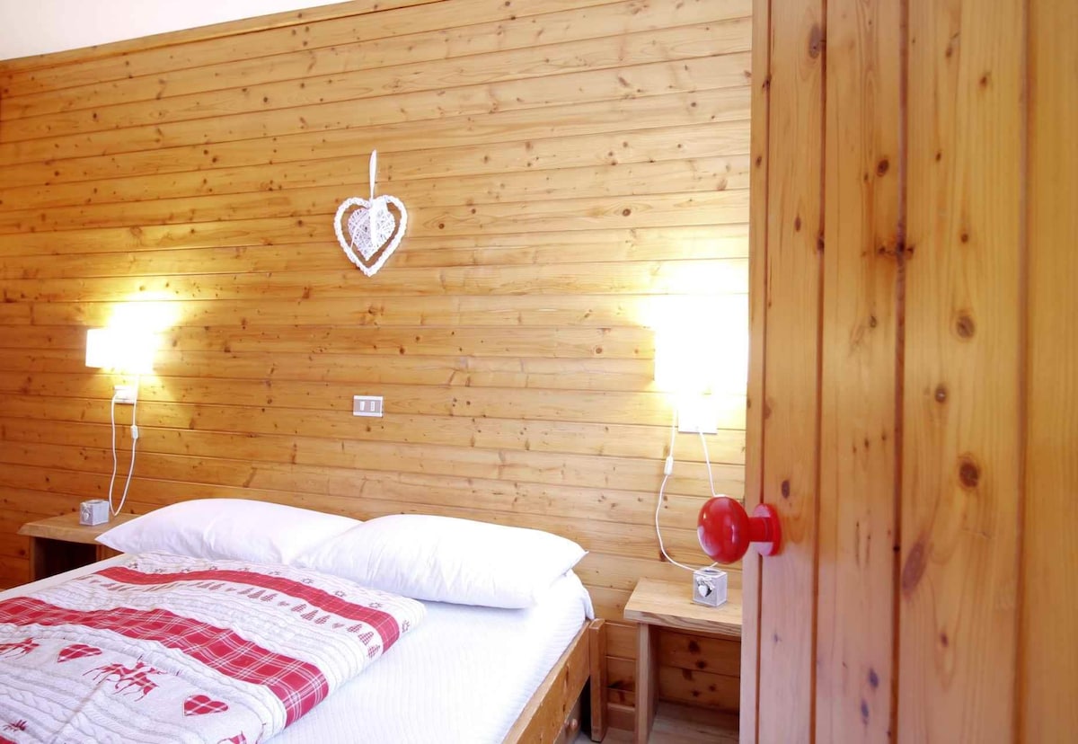 A cozy bedroom is presented with warm wood paneling. The spacious bed is adorned with a red and white patterned blanket. Bedside tables accompany the bed, each featuring a lamp. A heart-shaped decoration hangs on the wall, complementing the inviting atmosphere.